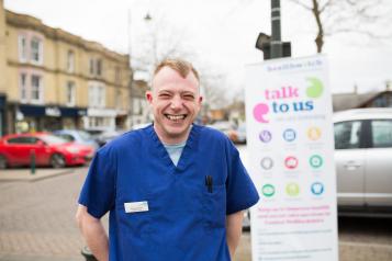 NHS worker standing infront of a Healthwatch sign that says 'talk to us'
